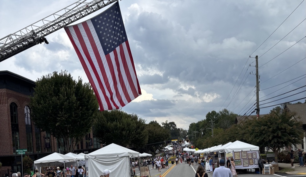 A large American flag hangs from a fire truck ladder over Crabapple Road, with white vendor tents and crowds stretching down the closed street under heavy gray clouds
