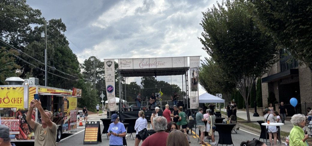 A band plays on the Crabapple Fest stage under gray clouds, with a food truck on the left and people at standing tables in the street